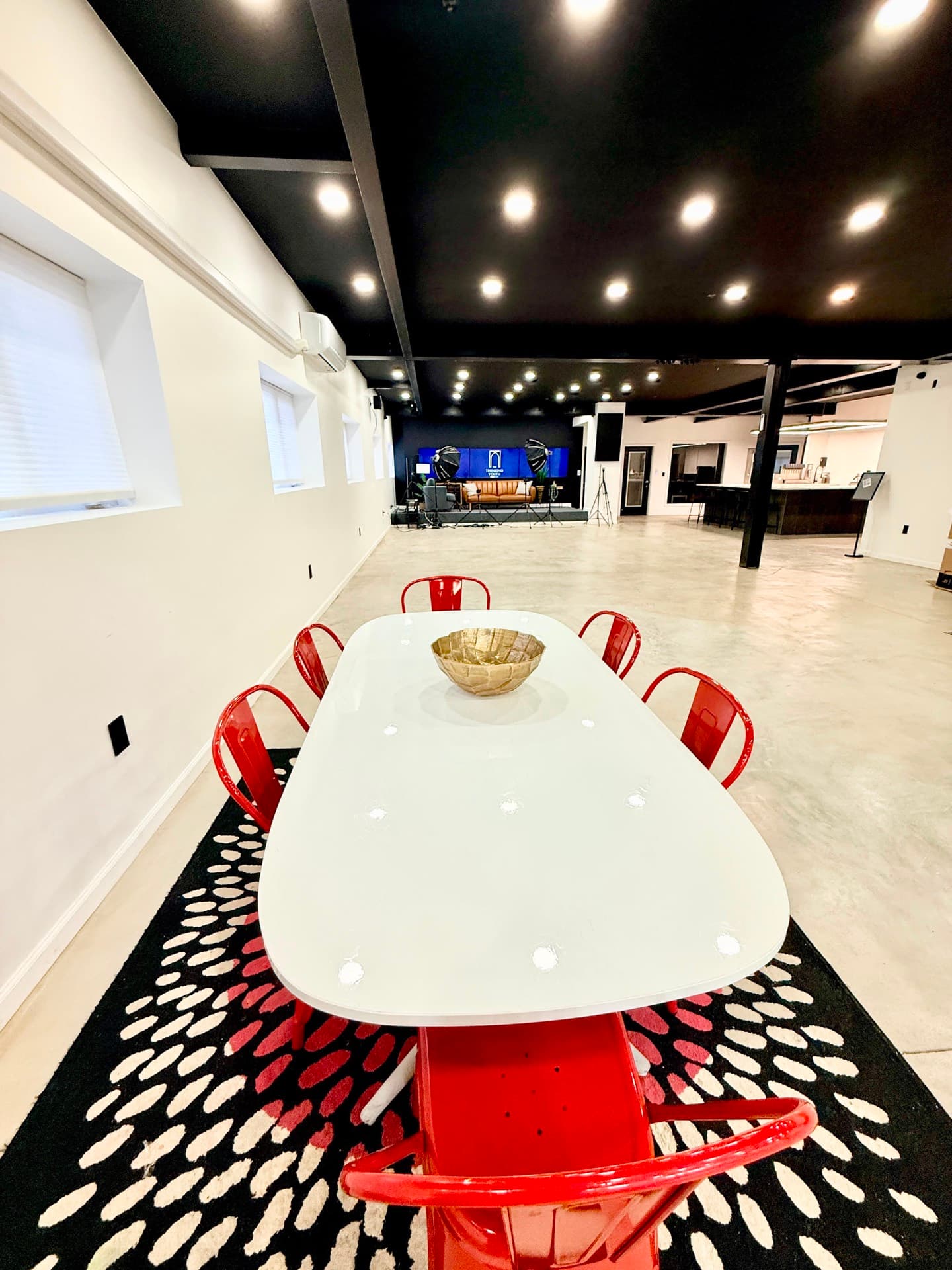 A long white table with red metal chairs down both sides, on a patterned black-and-white rug, in a room with white walls and high windows.